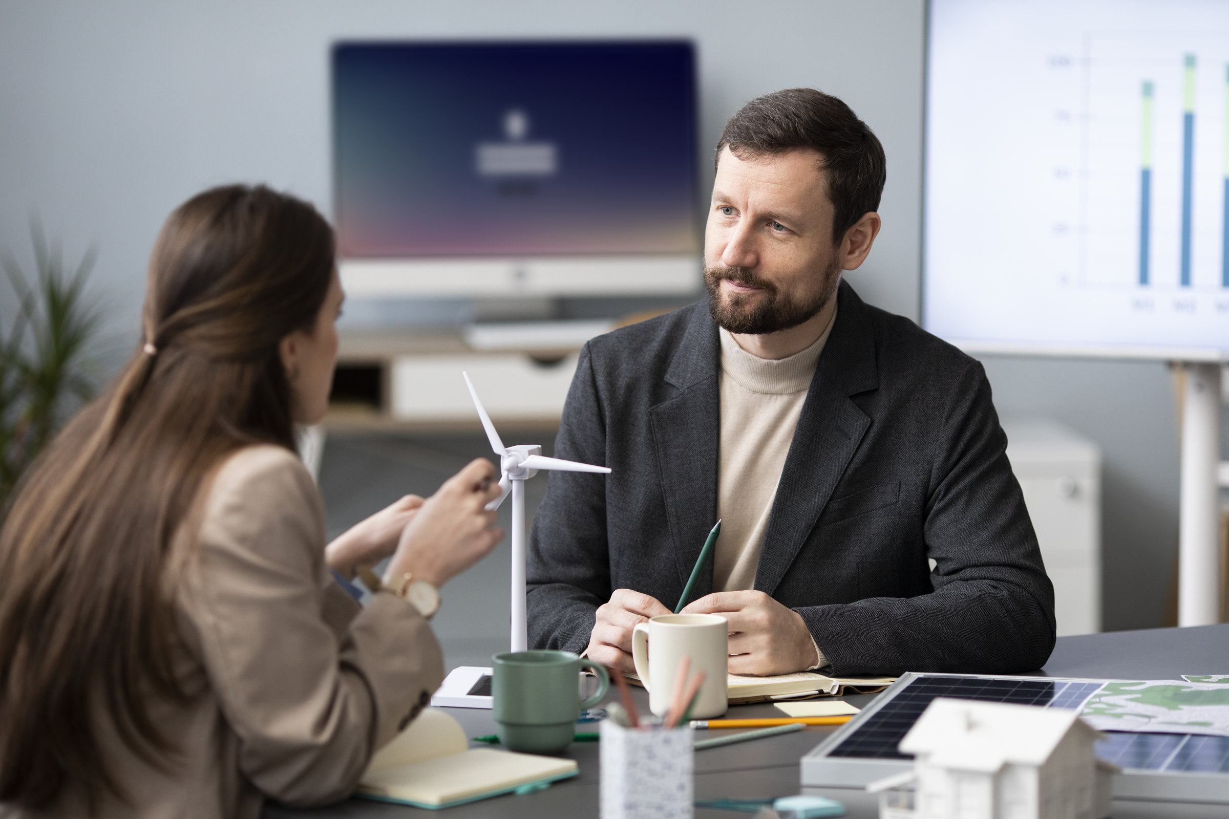 Two advisors discussing a project across a desk with a wind turbine model and a solar panel model